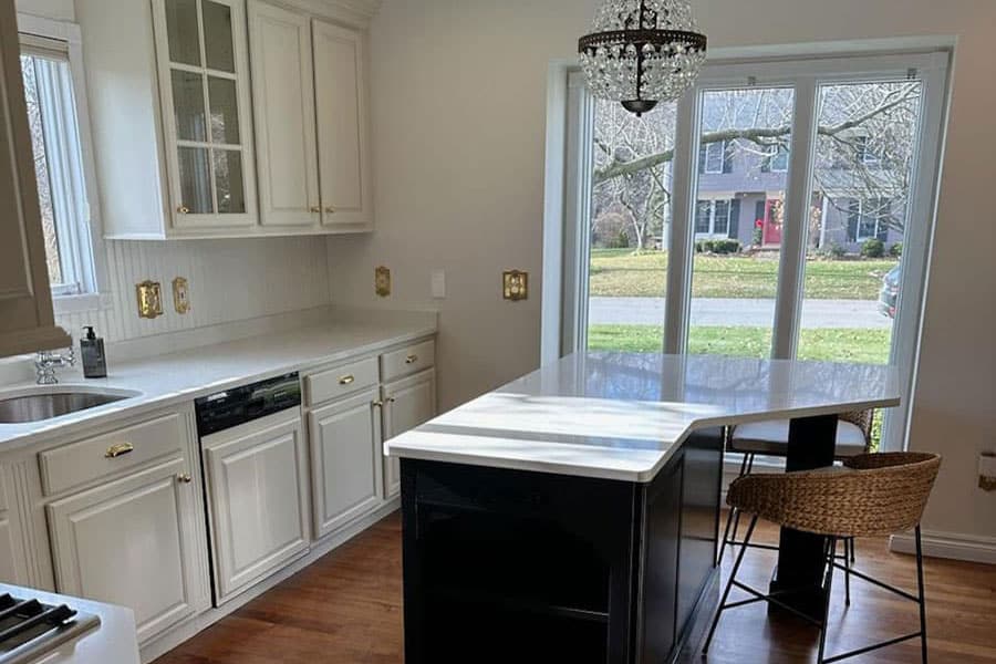 Bright kitchen with white cabinets featuring expert cabinet painting, marble countertops, a bold black island, wicker chairs, chandelier, and large windows overlooking a green yard and houses across the street in Royal Oak, MI.