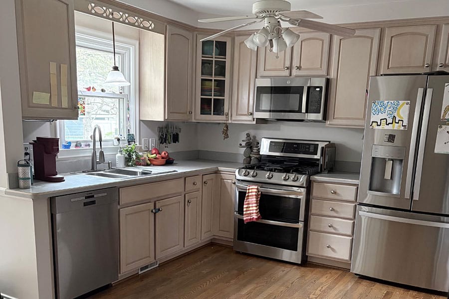 A modern kitchen with light wood cabinets, stainless steel appliances, a window over the sink, and a ceiling fan, this Harrison Township, MI home showcases the fresh look of professional cabinet painting. The fridge has kids art on it, and there’s a striped towel hanging on the oven.