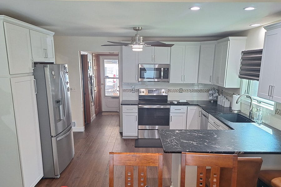 Modern kitchen in Royal Oak, MI with white cabinets, stainless steel appliances, black countertops, wooden floor, island with chairs, and a window with blinds letting in natural light. Ceiling fan and recessed lights are visible—an ideal space to consider Cabinet Refacing for a fresh update.