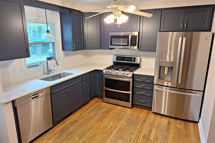 Modern kitchen with dark gray cabinets—perfectly suited for cabinet refinishing—stainless steel appliances, a gas stove, dishwasher, double-door fridge, white countertops, wood floor, and a ceiling fan with lights. The window above the sink offers a view of greenery outside, creating an inviting space in Metro Detroit.